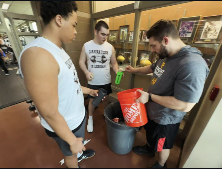 Varsity football players Chase Clark (left) and Declan Gill (center) receive pre workout powder from Coach Mark Oliver before a lift.